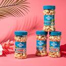 Four jars of Mauna Loa macadamia nuts with blue lids are arranged on a bright pink background, surrounded by tropical leaves and flowers, showcasing different flavors and packaging.